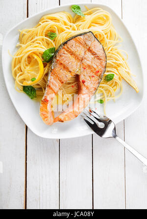 Meeresfrüchte Gourmetküche mit gegrilltem Lachs Schnitzel Steak serviert auf einem Bett aus italienischen Linguine Nudeln garniert mit Zitrone und frischem Basilikum, Draufsicht auf einen weißen Holztisch mit Exemplar Stockfoto