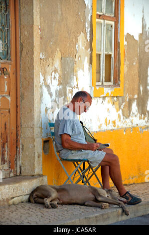 Algarve, Portugal, 24-September-2007: ein Mann sitzt auf einem Stuhl und las eine Zeitung mit seinem Hund neben ihm auf einer Straße in der Algarve. Stockfoto