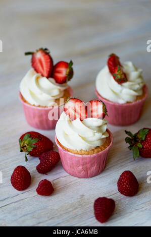 Muffins mit Sahne und süße Sahne, dekoriert mit Erdbeeren. Auf einem hölzernen Hintergrund. Stockfoto
