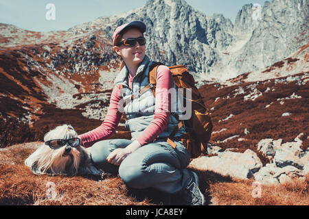 Touristen-junge Frau mit Hund und Sonnenbrillen sitzen auf hohen Bergen Hintergrund. Weißen und roten Herbstfarben. Stockfoto