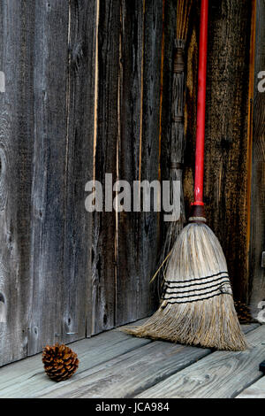 Alte Besen mit Orange-rötliche Griff rustikalen Veranda Wand mit Tannenzapfen im Vordergrund gelehnt. Stockfoto