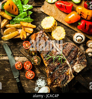 Blick von oben auf ein leckeres Gegrilltes Porterhouse Steak mit verschiedene gebratene Gemüse wie Tomaten, Paprika, Champignons, Mais, Zuckererbsen und Kartoffel wedges Stockfoto