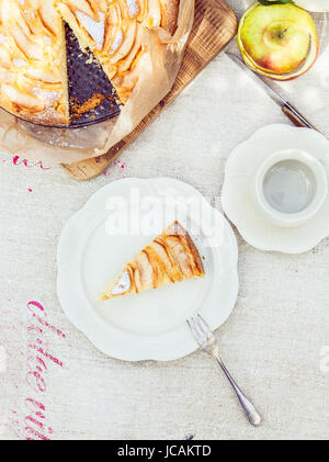 Stück leckeren frisch gebackenen Apfelkuchen serviert auf einem Teller mit einer leeren Tasse und Untertasse für Tee oder Kaffee neben den ganzen Kuchen auf ein weißes Tuch von oben gesehen Stockfoto