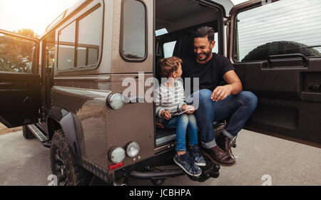 Schuss von Vater und Sohn sitzen auf der Rückseite des Autos und lächelnd. Mann mit kleiner Junge hält digital-Tablette während auf Roadtrip. Stockfoto