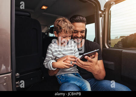 Vater und Sohn sitzen im hinteren Teil Auto und lächelnd mit digital-Tablette. Junger Mann mit kleinen Jungen halten digital-Tablette auf Roadtrip. Stockfoto