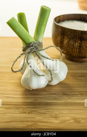 Drei Knoblauch Zwiebeln gefesselt zusammen mit Bindfäden auf Schneidebrett in der Nähe der Schüssel mit Salz. Unscharfen Hintergrund. Stockfoto