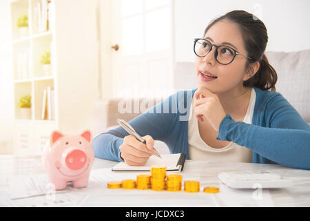 gerne asiatische chinesische Haus Frau träumen und Aufnahme schriftlich ihre Ersparnisse von Piggy Bank Geld Turm sitzt auf dem Sofa im Wohnzimmer zu Hause. IND Stockfoto