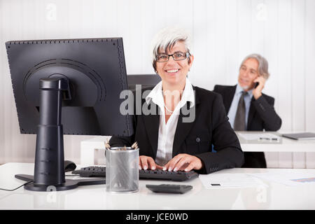 Lächelnde Sekretärin oder Assistentin sitzen an ihrem Schreibtisch arbeiten, mit ihrem Chef am Telefon im Hintergrund sichtbar Stockfoto