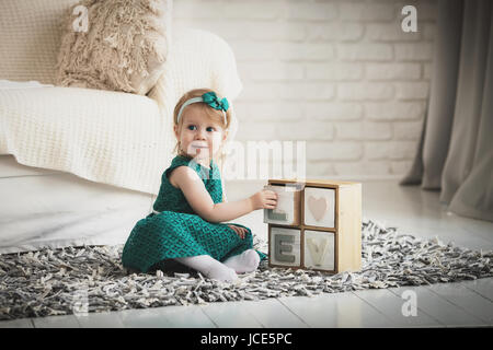 Kind-Modell mit Holzbuchstaben auf grauen flauschiger Teppich drinnen spielen. Kleines Mädchen im grünen Kleid in modernen Wohnung. Stockfoto