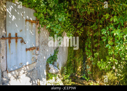 Ein Fenster mit geschlossenen alten, Metall Fensterläden auf einer Außenwand eines Hauses mit einer Wasserquelle auf der rechten Seite. Stockfoto