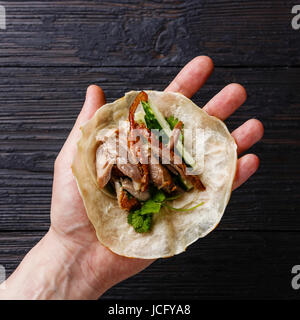 Peking-Ente mit Gurke, Frühlingszwiebeln und Koriander in gerösteten wheaten chinesischen Pfannkuchen in männlicher Hand Nahaufnahme auf schwarz verbrannten Holz Hintergrund Stockfoto