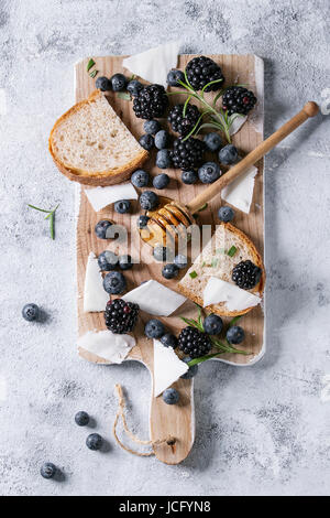 Beeren-Brombeere und Heidelbeere, Honig auf Dipper, Rosmarin, Ziegenkäse mit Brot serviert auf Holzbrett über graue Textur Hintergrund geschnitten. Sommer Stockfoto