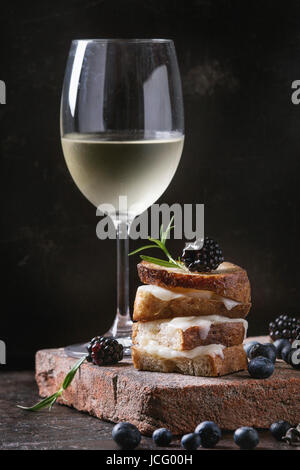 Gegrilltes Sandwich mit geschmolzenem Ziegenkäse, Brombeere, Heidelbeere, Rosmarin und Honig, serviert auf Terrakotta-Brett mit Glas kalten Weißwein über dar Stockfoto