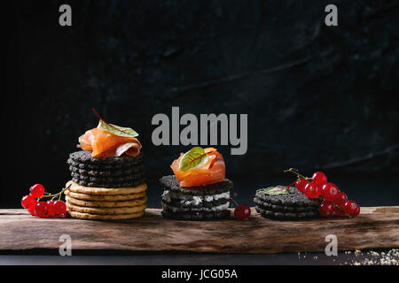 Stapel von schwarzen Holzkohle und traditionellen Cracker mit Räucherlachs, Frischkäse, grüner Salat und rote Johannisbeere Beeren auf Ood dienen Board über schwarz Stockfoto