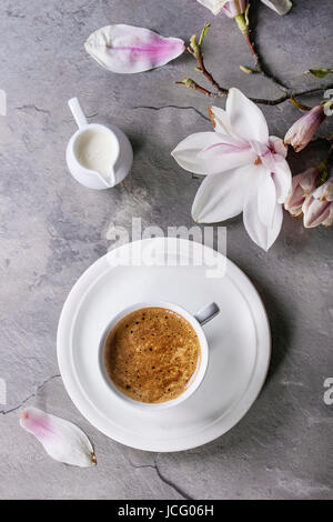 Weiße Tasse schwarzen Kaffee, serviert auf weiße Untertasse mit Krug mit Sahne und Magnolie Blume Blüte Zweig über graue Textur Hintergrund. Flache Lay, Raum Stockfoto