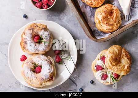 Füllen und leeren hausgemachte Choux Gebäck Kuchen Paris Brest mit Himbeeren, Mandeln, Puderzucker, Rosmarin auf Platte und Ofen Tablett mit Beeren über gra Stockfoto