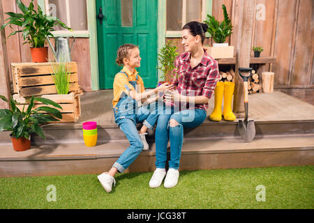 Glückliche Mutter und Tochter mit Topfpflanze zusammen auf der Veranda sitzen und Lächeln einander Stockfoto