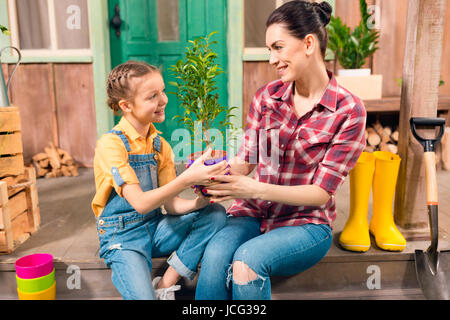 Glückliche Mutter und Tochter mit Topfpflanze zusammen auf der Veranda sitzen und Lächeln einander Stockfoto