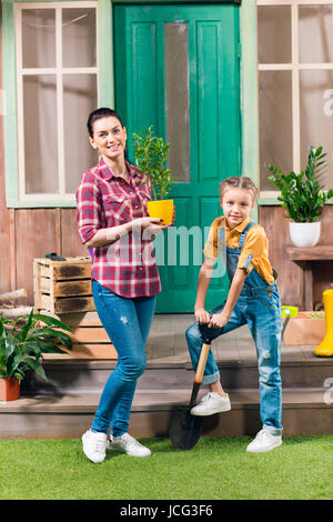 Lächelnde Mutter mit Topfpflanze und Tochter Garten Schaufel steht auf Veranda Stockfoto