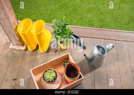 Gießkanne, gelbe Stiefel und Pflanzen in Holzkiste auf Veranda Stockfoto