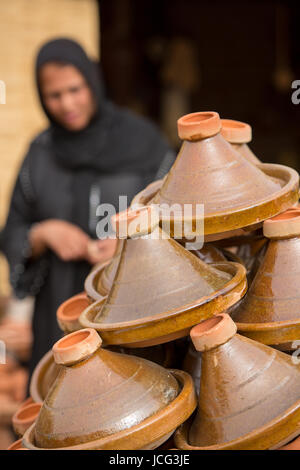 Auswahl an braunen marokkanische Tajines (traditionelle Auflaufformen) gestapelt auf dem Markt mit der Form einer unscharfen islamische Frau im Hintergrund Stockfoto