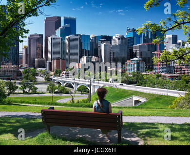 Junge Frau sitzt allein im Sunnyside Bank Park auf einer Bank an einem sonnigen Sommertag mit Centre Street Bridge und Calgary City Innenstadt schöne skylin Stockfoto