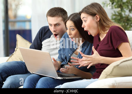 Drei Freunde, die Medieninhalte auf Linie in einem Computer sitzen auf einer Couch im Wohnzimmer zu Hause beobachten erstaunt Stockfoto