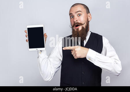 Gut aussehend Geschäftsmann mit Bart und Schnurrbart hält digital-Tablette und Blick auf Kamera und Bildschirm zeigt mit Finger und überrascht. Stud Stockfoto