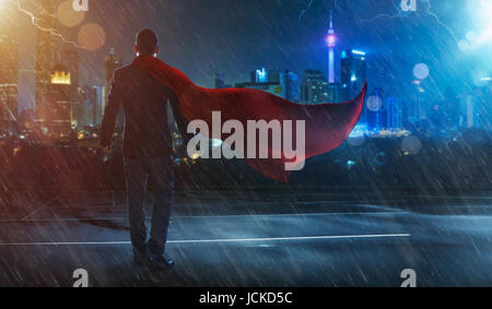 Geschäftsmann in Anzug und Cape Held vor einem Sturm, Sieg-Konzept. Stadtbild Hintergrund, Nachtaufnahme. Stockfoto