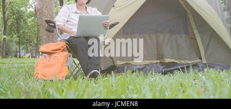 junge Asiatin Tourist auf Stuhl sitzend und mit Laptop vor Zelt am Campingplatz im Wald. Outdoor-Aktivitäten im Sommer. Travelin Abenteuer Stockfoto