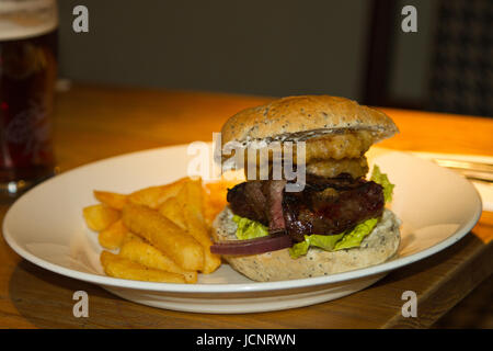 Traditionelle Steak Burger, Chips, Zwiebelringe mit einem Pint generische Bier oder Bitter im Hintergrund. Stockfoto