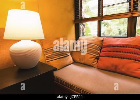Relax-Ecke der gelben Wand Haus mit bunten Kissen auf Bettsofa und helle große Lampe auf Tisch neben Sichtfenster durch Vorhang abgedeckt. Stockfoto