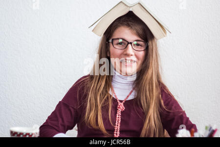 Schöne, niedliche kleine blonde Mädchen mit Brille und lila Kleid mit Buch auf dem Kopf überrascht und versuchen, etwas zu sagen Stockfoto