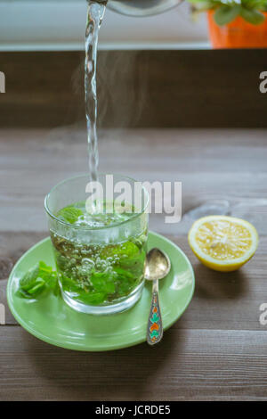 gießen heißes Wasser in einer transparenten Glastasse frischen Pfefferminztee auf einem dunklen Holztisch mit Blätter frische Minze und Zitrone Stockfoto