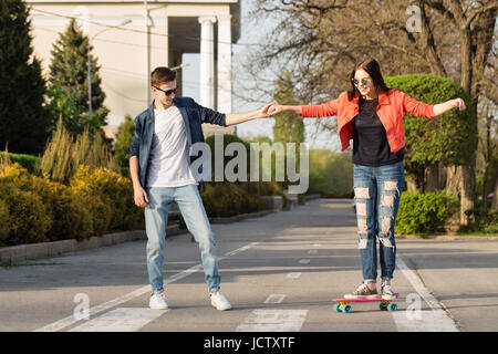 Verliebte Teenager. Der Kerl unterrichtet seine Freundin Skateboard. Datum der Hipster. Er hält ihre Hand. Romantik der ersten Liebe. Stockfoto