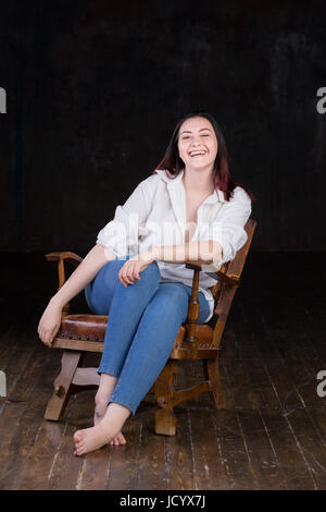 Schöne weiße braune kurzhaarige Hündin mit farbigen in roten enden tragen Männer weißes Hemd und blaue Jeans, sitzt auf einem Vintage Stuhl auf einem dunklen Hintergrund Stockfoto