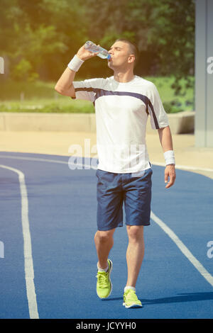 Porträt der durstige Sportler trinken Wasser aus der Plastikflasche und zu Fuß auf der blauen Laufbahn. Bäume und anderem Grün im Hintergrund Stockfoto