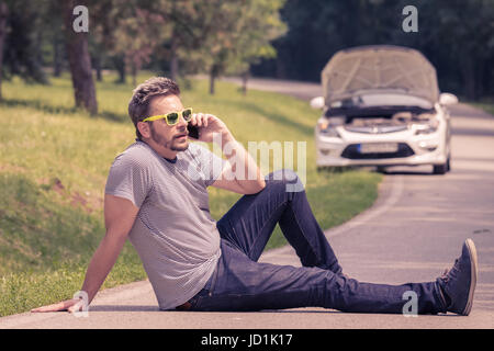 Junge moderne Mann sitzt auf den Asphalt und machen Anruf mit Hilfe Pannenhilfe. Auto mit angehobenen Haube am Straßenrand. Stockfoto