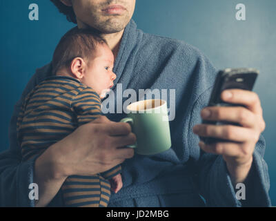 Ein junger Vater im Bademantel ist ein Smartphone verwenden, während Sie eine Tasse und sein kleiner Sohn festhalten Stockfoto