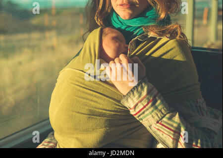 Am Bus ist eine junge Mutter mit ihrem Baby im Tragetuch bei Sonnenuntergang Stockfoto