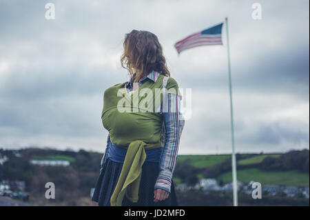 Eine junge alleinerziehende Mutter mit ihrem Baby im Tragetuch steht eine amerikanische Flagge Stockfoto