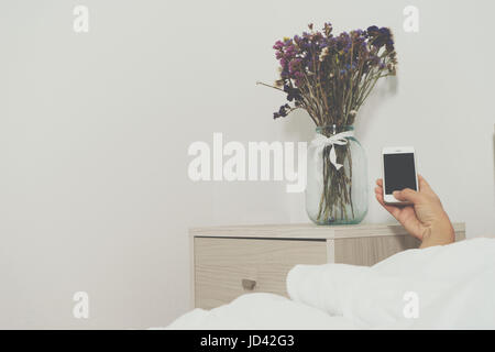 Womans Hand hält eine Telefon. Blume auf Tisch Bett in einem hellen Schlafzimmer Stockfoto