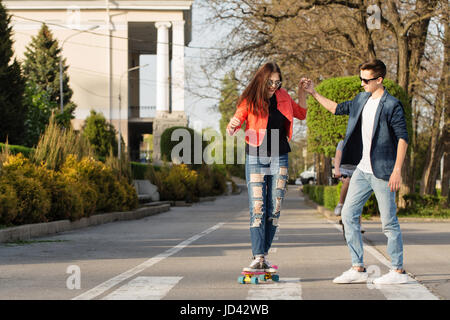 Verliebte Teenager. Der Kerl unterrichtet seine Freundin Skateboard. Datum der Hipster. Er hält ihre Hand. Romance erste Liebe. Stockfoto