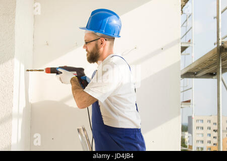 Bauarbeiter in Arbeit Kleidung, Schutzhandschuhe und einen Helm auf. bohren Sie das Loch mit einem Bohrer in der Wand. Stockfoto