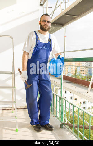 Bauarbeiter in einer Arbeit Kleidung, Schutzhandschuhe hält einen Helm und einen Hammer. Arbeiten in großer Höhe. Gerüst im Hintergrund Stockfoto