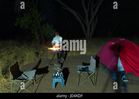 Frau, Kochen mit Feuer Holz und Braai Ausrüstung bei Nacht. Zelt und Stühle im Vordergrund. Abenteuer in afrikanischen Nationalparks. Stockfoto