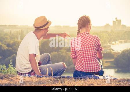Rückansicht des jungen Paares betrachten und genießen den Sonnenuntergang beim Sitzen auf der Wiese der Hügel über der Stadt. Sommer und Liebe Konzepte. Stockfoto