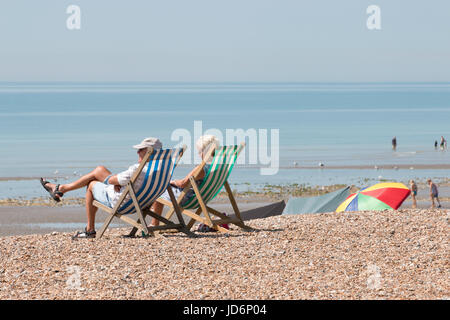 Ein paar sitzen auf Stühlen und die Sonne am Strand genießen in Worthing. Die Flut ist, und die Menschen sind das Paddeln im Hintergrund. Stockfoto