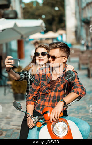 Junges attraktives paar Teenager trägt Sonnenbrille auf Retro-Motorrad macht Selfie, sonnige Straße, urban Hipster-Konzept Stockfoto