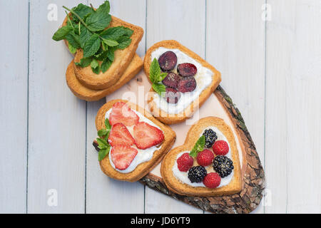 Herzförmige Kekse verteilt mit Quark, Erdbeeren, Brombeeren, Himbeeren, Kirschen und einem Zweig Minze präsentiert auf einem Baum Datenträger Stockfoto